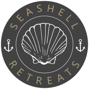 Rundes Logo von Seashell Retreats mit stilisierter Muschel in der Mitte und zwei Ankern auf dunklem Hintergrund.