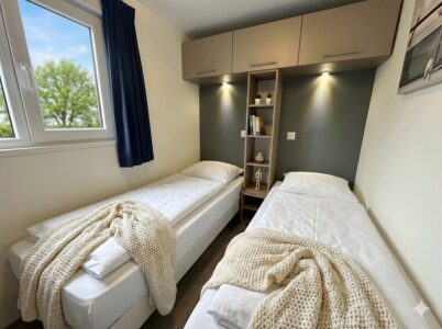 Helles Schlafzimmer mit zwei Einzelbetten, Regal und Leselampen, Fenster mit Vorhang und Blick ins Grüne. - Villa Sunset Beach